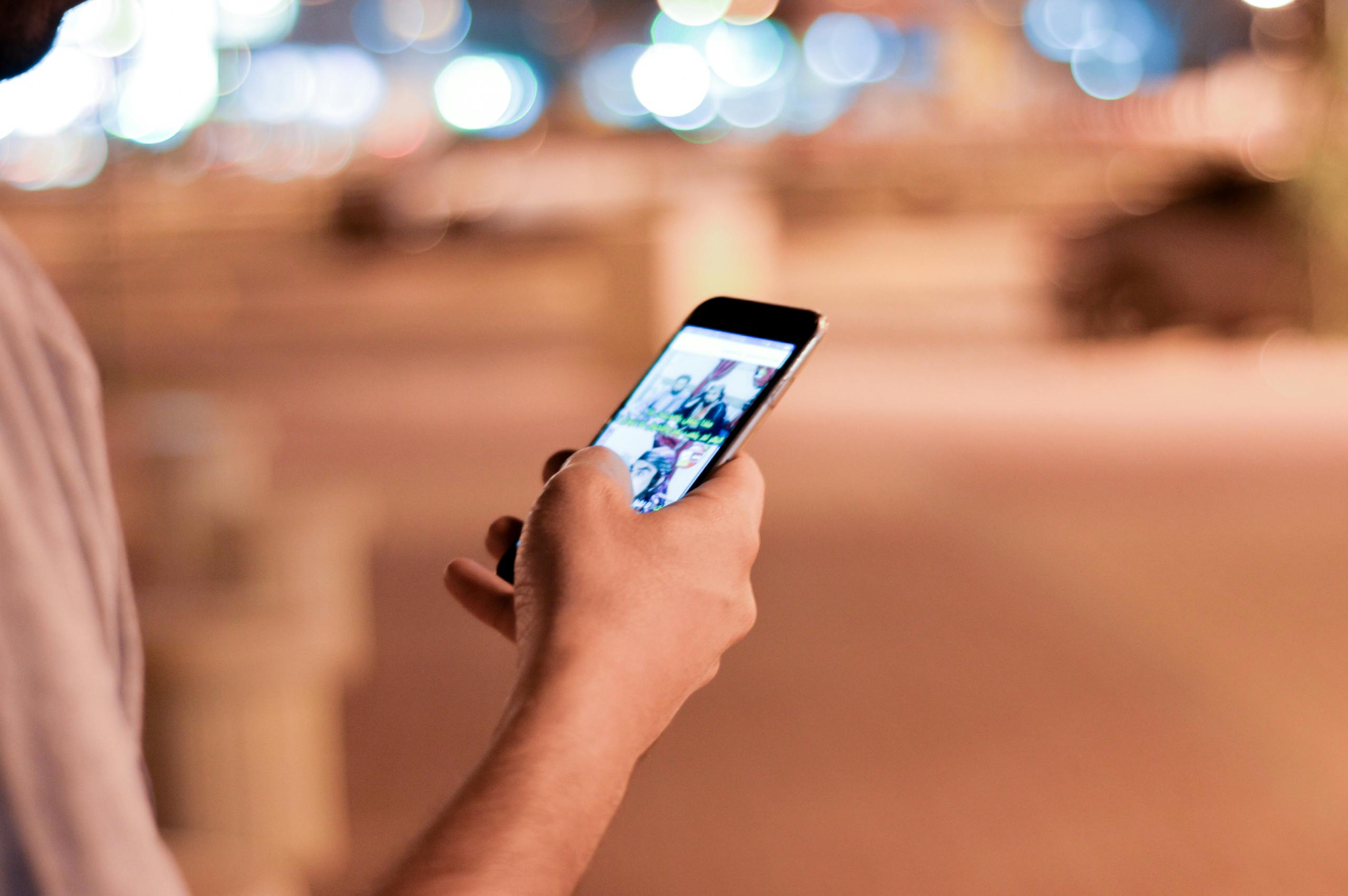 A close-up of a person using a smartphone outdoors at night, with blurred bokeh lights in the background.